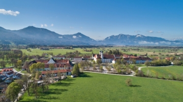 Luftaufnahme des Klosters Benediktbeuern vor malerischer Alpenkulisse, eingebettet in grüne Wiesen und weitläufige Landschaft.