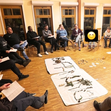 In einem erleuchteten Raum sitzen Erwachsene und Jugendliche in einem Stuhlkreis. Auf dem Parekttboden sind zwei Weiße rechteckige Decken mit Blättern, Zweigen und Steinen.