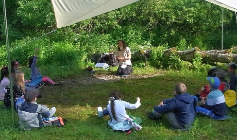 Kindergruppe mit Anleiterin lernt auf einer Wiese unter einem aufgespannten Segeltuch
