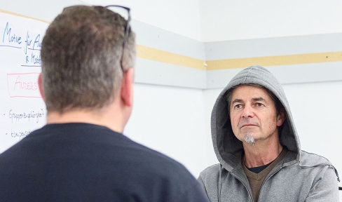 Ein Mann im Kapuzenpulli steht mit verschränkten Armen einem anderen Mann gegenüber, der nur von hinten zu sehen ist.
